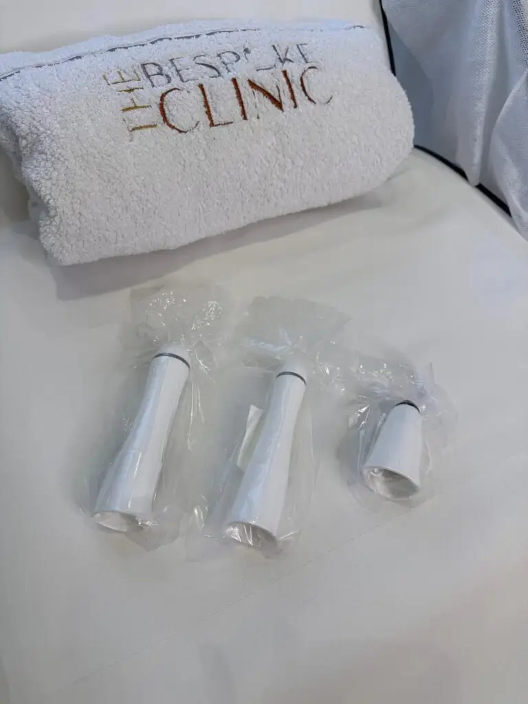 Rolled white towel with brown embroidery resting on a surface; in front are several small white plastic tubes sealed in plastic bags.