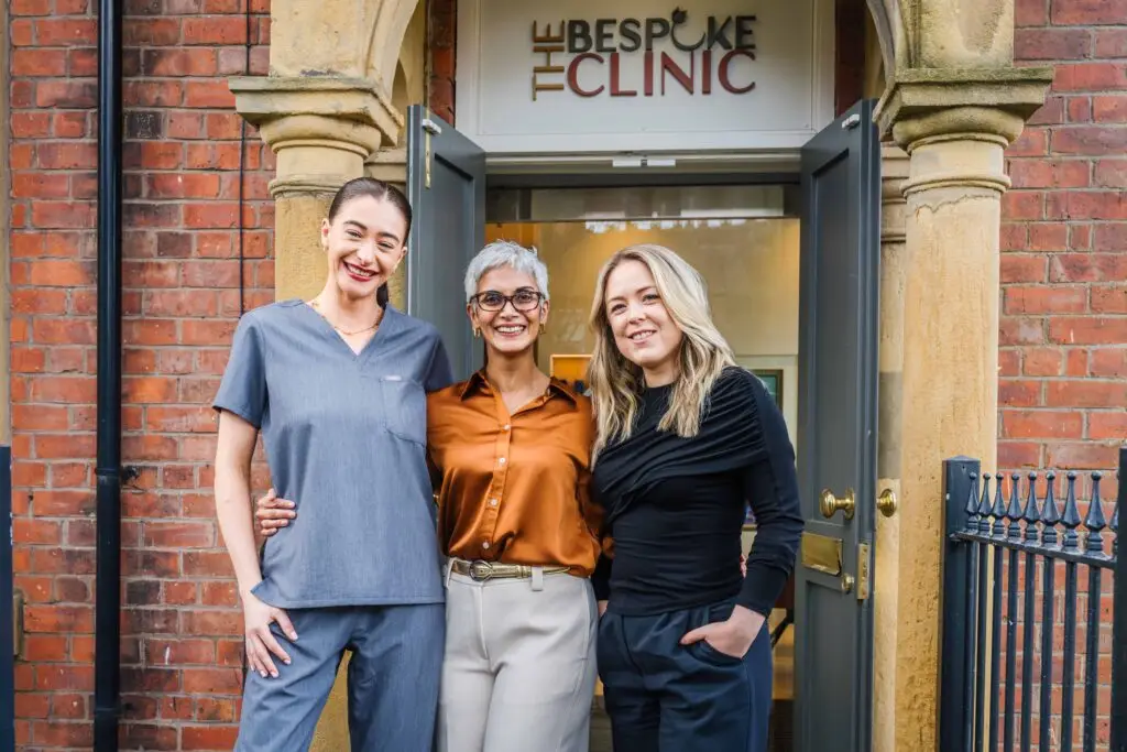 Three women stand together outside a brick building with an arched doorway that reads 'The Bespoke Clinic,' smiling at the camera.